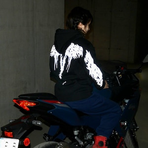 May include: A black hooded sweatshirt featuring a white graphic design of wings on the back and sleeves. The hoodie is worn with dark blue pants while seated on a dark blue motorcycle with a visible red taillight and a white license plate.
