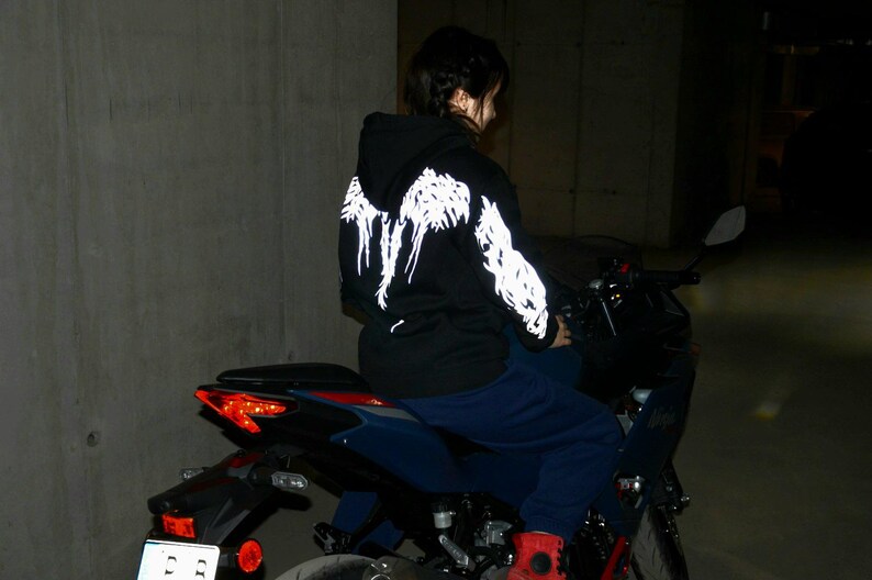 May include: A black hooded sweatshirt featuring a white graphic design of wings on the back and sleeves. The hoodie is worn with dark blue pants while seated on a dark blue motorcycle with a visible red taillight and a white license plate.