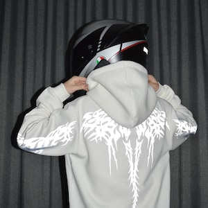 May include: A light gray hooded sweatshirt featuring a reflective white graphic print on the back and sleeves, paired with a black and silver motorcycle helmet. The hoodie has long sleeves and a hood that rests against the back of the helmet.