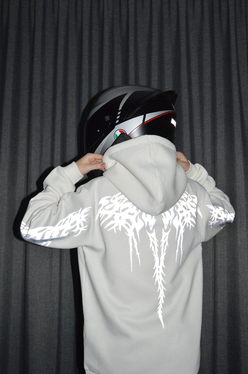 May include: A light gray hooded sweatshirt featuring a reflective white graphic print on the back and sleeves, paired with a black and silver motorcycle helmet. The hoodie has long sleeves and a hood that rests against the back of the helmet.
