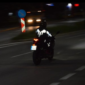 May include: A motorcyclist wearing a black jacket with a reflective white wing design on the back, riding a motorcycle at night. The motorcycle features a red taillight and a license plate with the text P B 3436, visible on a dark road.