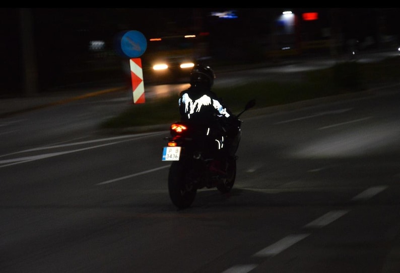 May include: A motorcyclist wearing a black jacket with a reflective white wing design on the back, riding a motorcycle at night. The motorcycle features a red taillight and a license plate with the text P B 3436, visible on a dark road.