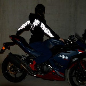 May include: A black hooded sweatshirt featuring reflective white wing graphics along the sleeves and back, worn while sitting on a dark blue and red Kawasaki Ninja motorcycle with visible branding on the side fairing and a bright front headlight.