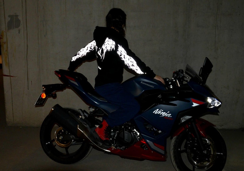 May include: A black hooded sweatshirt featuring reflective white wing graphics along the sleeves and back, worn while sitting on a dark blue and red Kawasaki Ninja motorcycle with visible branding on the side fairing and a bright front headlight.