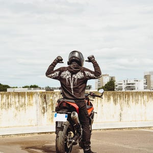 May include: A person wearing a black hooded sweatshirt with a white skeletal wing graphic on the back, black pants, black gloves, and a black helmet, sitting on a red and black motorcycle. The motorcycle has a visible rear tire, exhaust, and license plate.