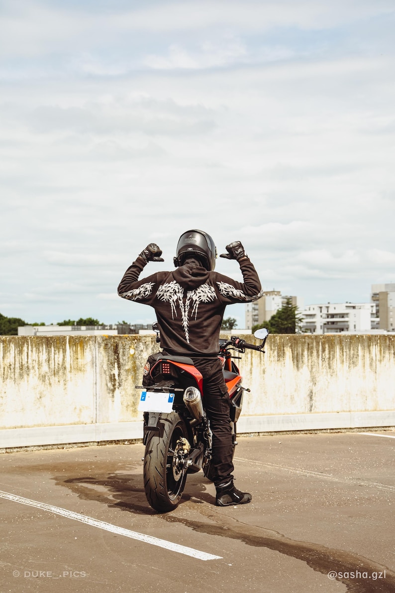 May include: A person wearing a black hooded sweatshirt with a white skeletal wing graphic on the back, black pants, black gloves, and a black helmet, sitting on a red and black motorcycle. The motorcycle has a visible rear tire, exhaust, and license plate.