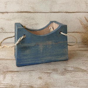 May include: A rectangular wooden crate with a distressed blue finish and a curved top edge. The container features two natural fiber rope handles attached to the sides, with the interior showing the natural light brown wood grain texture.