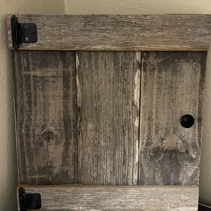 May include: A rustic wooden cabinet with a weathered gray finish. The cabinet features a black knob, black hinges, and is constructed from reclaimed wood. The design is simple and the wood grain is visible.
