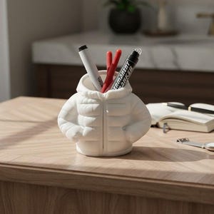 May include: A white ceramic pen holder shaped like a puffy jacket, holding a white pen, two red pens, and a black marker. The pen holder is on a wooden table, with a notebook, watch, and coffee cup in the background.