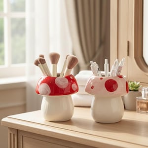 Puede incluir: Dos recipientes de cerámica con forma de hongo, uno rojo con lunares blancos y otro rosa con lunares rojos, contienen brochas de maquillaje y bolígrafos. Los recipientes tienen una base blanca y están sobre una superficie de madera.