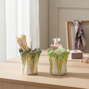 Op de afbeelding: Twee keramische containers in de vorm van koolbladeren, waarvan één met make-up borstels. De containers zijn wit met groene en gele accenten en staan op een licht houten oppervlak. De achtergrond omvat een raam, een fotolijst en een parfumfles.