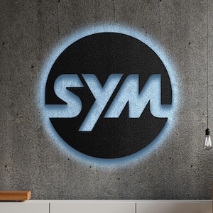 May include: The SYM logo is displayed on a textured, gray concrete wall. The logo features the letters "SYM" in a light blue color, set against a black circle with a blue outline. A light bulb hangs in the background.