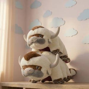 May include: Two plush Appa figures from Avatar: The Last Airbender, one stacked on top of the other. The figures are primarily white with brown patches, gray muzzles, and curved horns. They sit on a wooden surface against a pink wall with cloud cutouts.