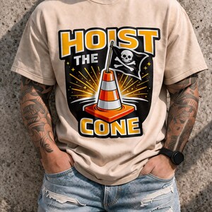May include: Beige t-shirt with a graphic featuring a traffic cone, a pirate flag with a skull and crossbones, and the words "HOIST THE CONE" in yellow and orange. The design has a black background with starburst details.