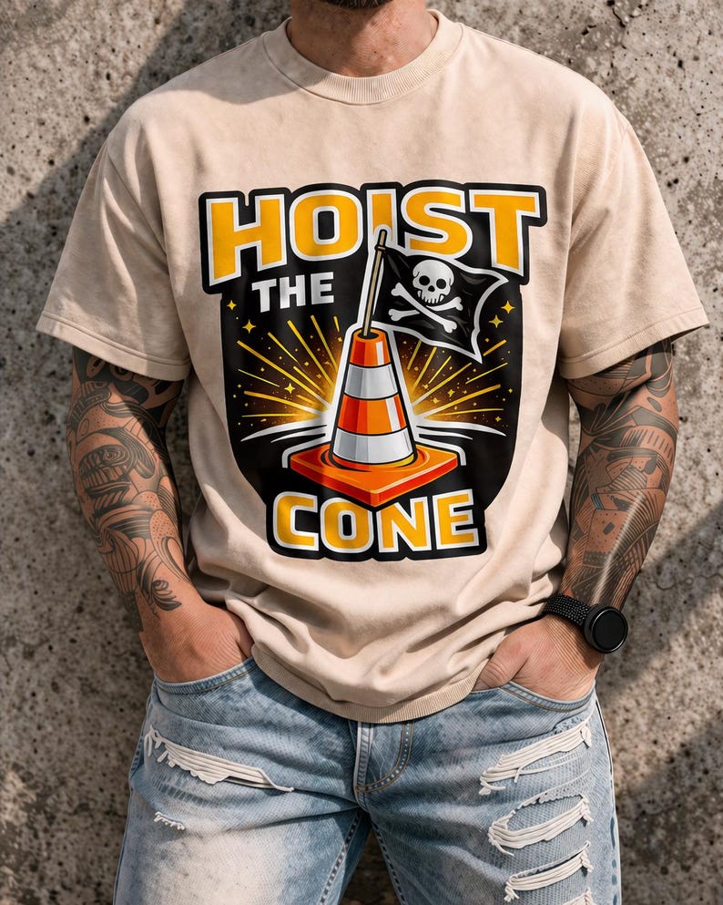 May include: Beige t-shirt with a graphic featuring a traffic cone, a pirate flag with a skull and crossbones, and the words "HOIST THE CONE" in yellow and orange. The design has a black background with starburst details.