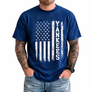 Peut inclure: T-shirt bleu marine avec un motif de drapeau américain vieilli et le mot "Yankees" en texte vertical blanc. Le t-shirt a un col rond et des manches courtes. Le motif incorpore des étoiles et des rayures, représentant les États-Unis.
