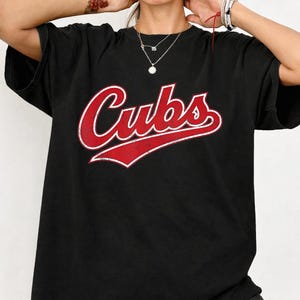 Peut inclure: T-shirt noir avec le mot "Cubs" en rouge et blanc. Le mot "Cubs" est dans une police stylisée avec un contour rouge et un remplissage blanc. Le t-shirt est de style décontracté.
