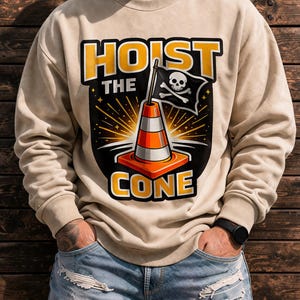May include: Beige sweatshirt with the words "HOIST THE CONE" in yellow and black. The graphic features a traffic cone with a skull and crossbones flag. The sweatshirt is worn with blue jeans.