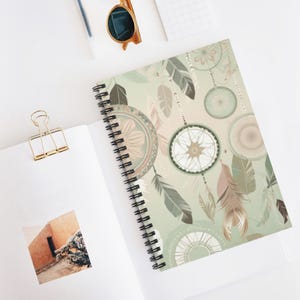 May include: Spiral-bound notebook with a cover featuring a dreamcatcher design in shades of green, beige, and brown. The design includes dreamcatchers and feathers. The notebook is open, revealing blank pages.