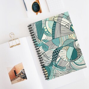 Könnte beinhalten: Ein spiralgebundenes Notizbuch mit einem farbenfrohen abstrakten Design in Türkis, Blau und Creme. Das Cover zeigt ein Muster aus Linien, Formen und Punkten. Das Notizbuch liegt offen auf einer weißen Oberfläche, mit einer goldenen Büroklammer und Sonnenbrille.