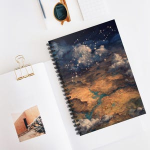 May include: Spiral-bound notebook featuring a celestial design. The cover displays a detailed illustration of a night sky with constellations, clouds, and a landscape in shades of blue, brown, and gold. The notebook is open on a white surface.