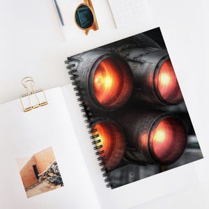 May include: Spiral-bound notebook featuring a detailed image of four jet engine nozzles with glowing orange and red interiors. The notebook is open on a white surface, with a gold paper clip and sunglasses visible.