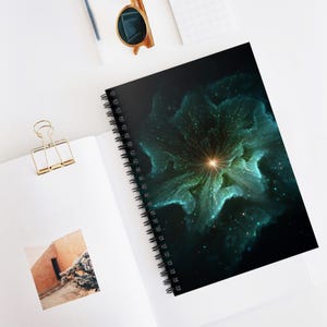 Könnte beinhalten: Spiralgebundenes Notizbuch mit einem abstrakten Blumendesign in Türkis und Gold. Das Cover zeigt ein leuchtendes Zentrum mit strahlenförmigen Linien vor dunklem Hintergrund. Das Notizbuch liegt offen auf einer weißen Oberfläche.