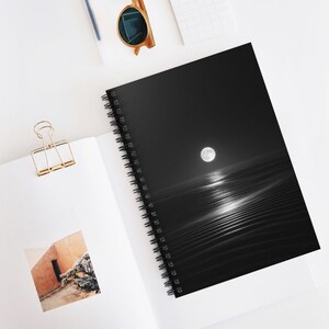 May include: Spiral-bound notebook featuring a black and white photograph of a full moon over rippled sand dunes. The image is printed on the cover, with a bright moon reflecting on the water. The notebook is open on a white surface.