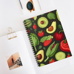 May include: Spiral-bound notebook with a black cover displaying a vibrant pattern of vegetables. The design includes illustrations of avocados, tomatoes, bell peppers, lettuce, and cucumbers. The notebook is open, revealing blank white pages.