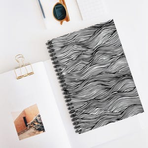 Puede incluir: Un cuaderno de espiral con una cubierta con un patrón de ondas en blanco y negro. El cuaderno está abierto, revelando páginas blancas en blanco. También se ven un clip dorado y gafas de sol.