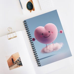 May include: Spiral-bound notebook featuring a whimsical illustration of a pink heart character with a smiling face, arms, and legs, floating on a cloud against a light blue background. The notebook is open, revealing a blank page and a gold binder clip.