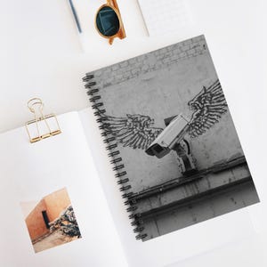 Könnte beinhalten: Spiralgebundenes Notizbuch mit einem Schwarz-Weiß-Bild einer Überwachungskamera mit Graffiti-Flügeln. Das Kunstwerk befindet sich auf einer grauen Wand mit Ziegeldetails. Das Notizbuch ist geöffnet und zeigt eine leere Seite und eine goldene Büroklammer.