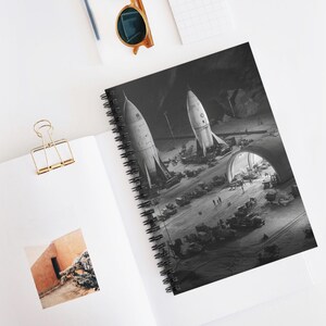 May include: Spiral-bound notebook featuring a black and white illustration of two rockets on a launchpad. The image includes a tunnel, vehicles, and figures, evoking a retro space exploration theme. The notebook is open on a white surface.