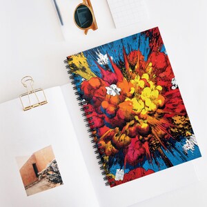 May include: Spiral-bound notebook featuring a vibrant comic book-style explosion design. The cover showcases a burst of red, orange, yellow, and blue colors, with a black spiral binding. The notebook is open on a white surface.