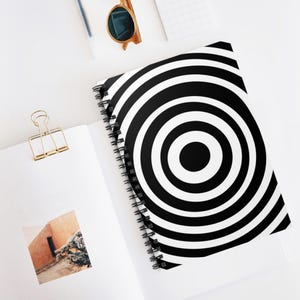 Puede incluir: Un cuaderno con espiral con un diseño de círculos concéntricos en blanco y negro. El cuaderno está abierto, revelando páginas blancas en blanco. También se ven un clip dorado y gafas de sol.