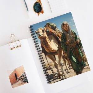 May include: Spiral-bound notebook featuring a vibrant illustration of a person in traditional attire leading a camel through a desert landscape. The camel is laden with colorful packs, and the background shows a clear blue sky.