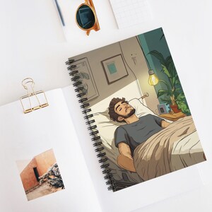 May include: Spiral-bound notebook with an illustration of a person asleep in bed. The artwork shows a bedroom scene with a hanging light, plants, and framed pictures. The person is wearing a grey shirt and is covered with a beige blanket.
