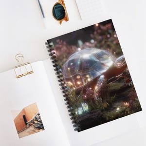 Puede incluir: Cuaderno de espiral con una ilustración detallada de una tortuga con un caparazón brillante e iridiscente. La tortuga está situada en un jardín exuberante y colorido con iluminación suave. El cuaderno está abierto sobre una superficie blanca con un clip dorado y gafas de sol.