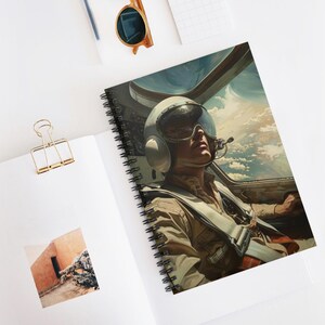 Può includere: Quaderno a spirale con un'illustrazione di un pilota in una cabina di pilotaggio, con un casco con visiera. L'opera d'arte ha uno stile vintage, incentrato sul volto del pilota e sul cielo. Il quaderno è aperto su una superficie bianca.