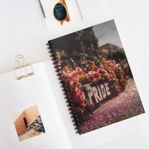 Può includere: Quaderno a spirale con una copertina raffigurante una fioriera piena di fiori con la scritta "PRIDE". Glitter rosa e una bandiera arcobaleno aggiungono alla scena. Il quaderno è aperto su una superficie bianca.