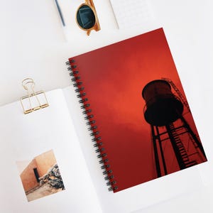 May include: Spiral-bound notebook with a vibrant red cover featuring a silhouette of a water tower against a dramatic sky. The notebook is open, revealing a blank page and a gold binder clip.