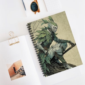 Puede incluir: Cuaderno de espiral con una ilustración de fantasía de una figura humanoide adornada con hojas verdes y armadura. La obra de arte tiene una paleta de colores apagados con tonos de verde, gris y beige. El cuaderno está abierto, revelando la ilustración.