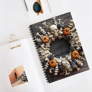 May include: A spiral-bound notebook with a Halloween wreath design. The wreath includes skulls, bones, orange pumpkins, and pinecones, set against a dark wood background. The notebook is open on a white surface, with a gold binder clip and sunglasses visible.