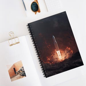 May include: Spiral-bound notebook featuring a dramatic image of a rocket launching into space. The cover art depicts the rocket ascending through a fiery atmosphere, with vibrant orange and red hues. The notebook is open, revealing blank pages.