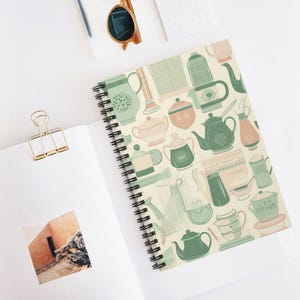 Pode incluir: Caderno espiralado creme com capa estampada com bules e chávenas em tons de verde e pêssego. O caderno está aberto, revelando páginas em branco e uma pequena foto. Óculos de sol e um clip também são visíveis.