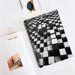 May include: A spiral-bound notebook with a black and white 3D cube design. The notebook is open, revealing blank white pages. A gold binder clip, sunglasses, and a pen are also visible.