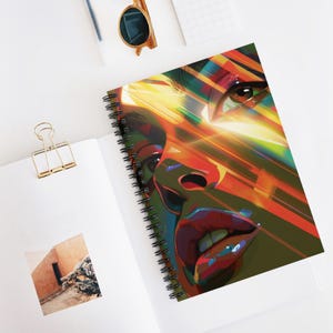 May include: Spiral-bound notebook featuring a vibrant, abstract portrait of a person's face. The artwork uses bold colors like red, yellow, and green, with a focus on the eyes and lips. The notebook is open on a white surface.