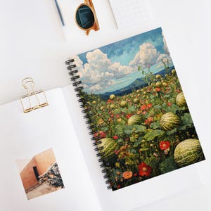 Puede incluir: Cuaderno de espiral con una colorida ilustración de un campo de sandías y flores bajo un cielo azul con nubes blancas. El cuaderno está abierto sobre una superficie blanca, con un clip dorado y gafas de sol.