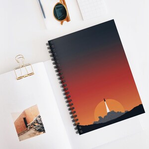 May include: Spiral-bound notebook featuring a vibrant sunset design with a rocket launching into the sky. The cover transitions from a deep navy blue to a fiery red and orange, with a silhouette of a mountain range and a large sun.