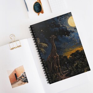 May include: Spiral-bound notebook featuring a giraffe under a starry night sky with a full moon. The cover art depicts a giraffe standing in a forest, with a dark blue and yellow sky. The notebook is open on a white surface.